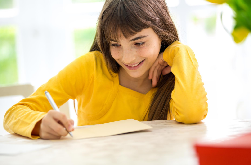 Late elementary or early middle school child wearing a bright yellow shirt writing a letter at a sunny spot in the kitchen. 