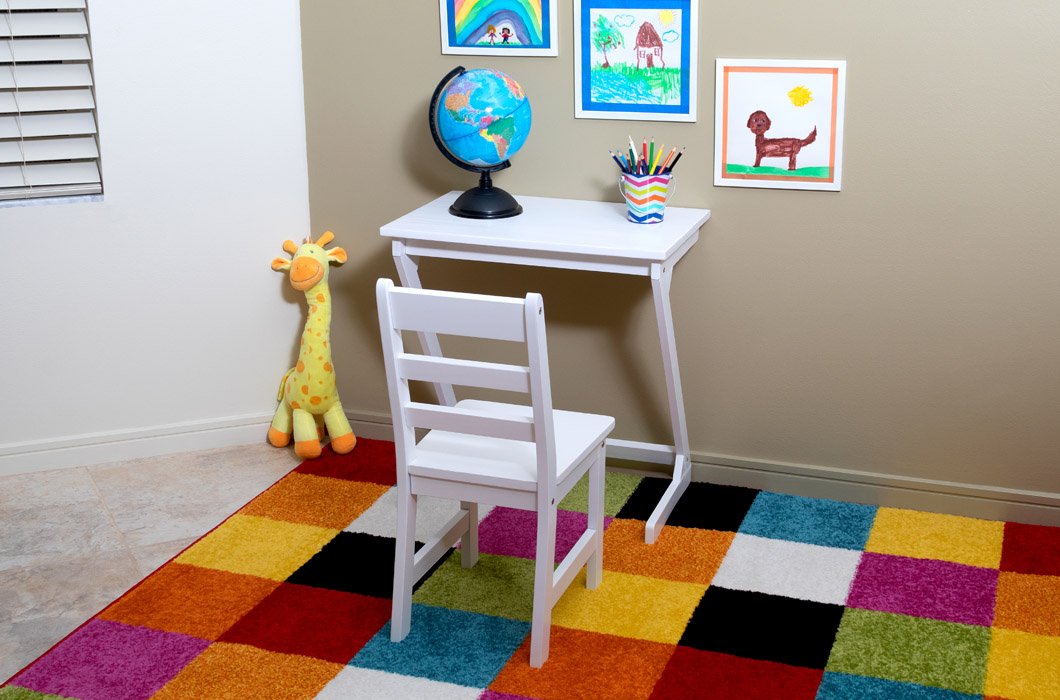 Lipper International Item 547W, The Child's Anywhere Table and Chair set, on a brightly colored checkered rug next to a toy giraffe. There is a small globe and a colored pencil cup on the surface of the table and there are children's drawings on the wall. 